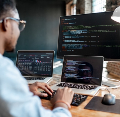 Young african male programmer writing program code sitting at the workplace with three monitors in the office. Image focused on the screen