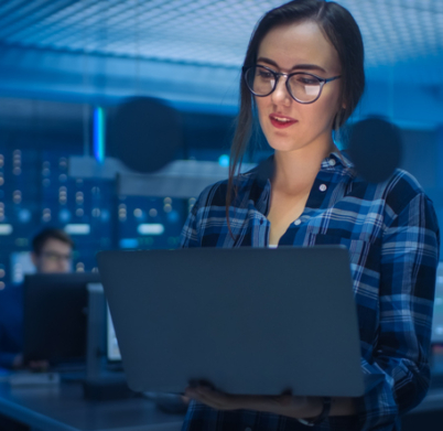 Portrait of a Smart Young Woman Wearing Glasses Holds Laptop. In the Background Technical Department Office with Specialists Working and Functional Data Server Racks